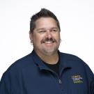 Smiling man with dark hair, mustache and goatee, navy shirt with yellow logo, white background